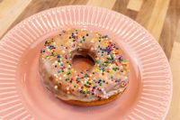 A glazed donut from the Donuts and Donut Holes recipe, covered in colorful sprinkles, on a pink plate.