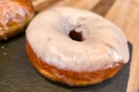 A glazed doughnut from Donuts and Donut Holes resting on dark slate, with a wooden board behind it.
