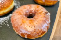 Close-up of a glazed doughnut from Donuts and Donut Holes on a dark slate surface, with two more doughnuts partly visible.