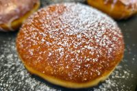 Close-up of a Czech Filled Donuts Koblihy dusted with powdered sugar, more donuts blurred behind it.
