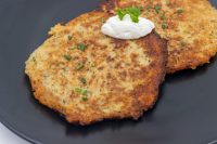 Two crispy Potato Pancakes Bramboraky on a dark plate, topped with sour cream and chopped scallion.