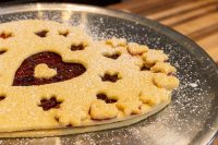 Linzer Tart Hearts with a heart-shaped cutout showing red preserve, edged in flower cutouts, dusted with sugar.
