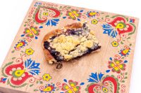 A slice of Sheet Kolach with Streusel showing dark berry filling and buttery crumb topping on a painted wooden board.