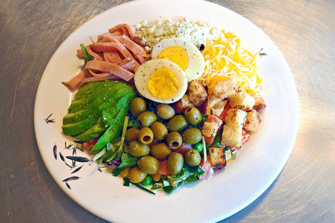 Cobb Salad Cobb Salad on a white plate with ham, egg halves, cheeses, avocado, olives, and croutons over greens.