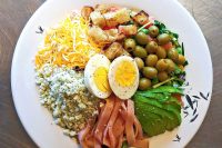 Cobb Salad Cobb Salad on a white plate, sectioned with cheeses, egg, olives, avocado, ham strips, and croutons.