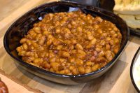 Dark square bowl filled with baked beans coated in thick brown sauce, with bread visible at the edge.