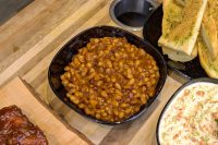 Bowl of Baked Beans in a rich brown sauce, served on a wooden board with garlic bread and coleslaw nearby.