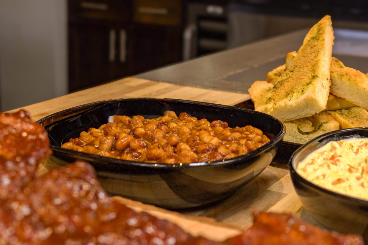 Black dish of Baked Beans in reddish sauce, served with garlic bread, a creamy side, and ribs.