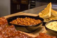 Black dish of Baked Beans in reddish sauce, served with garlic bread, a creamy side, and ribs.