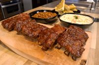 Glazed American BBQ – Honey Pork Ribs on a wooden board, with baked beans, coleslaw, and bread slices behind