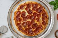Overhead shot of 60 min. Pan Pizza with pepperoni and melted cheese on a metal pan, basil nearby.